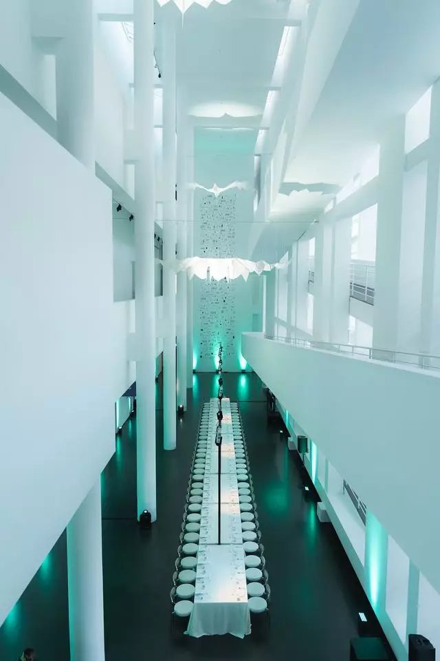 Una taula llarga parada per sopar a l'atri blanc del MACBA, il·luminada en turquesa